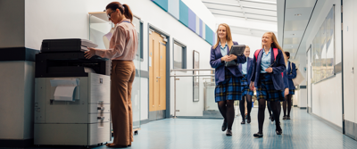 School teacher using a photocopier, children in corridor, smiling